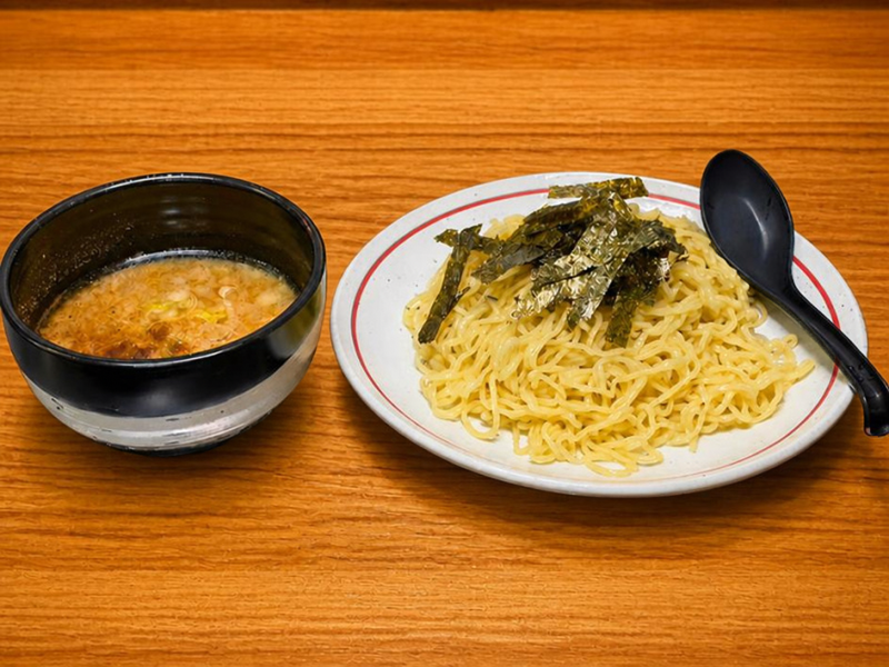 つけ麺処Sin 富士本店のメインメニュー「豚骨つけ麺」。木目のテーブルに置かれた、ネギが浮かぶ濃厚な豚骨スープが入った黒い器と、海苔がトッピングされた麺。