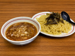 富士市でつけ麺・ラーメンを提供する「つけ麺処Sin 富士本店」の定番メニュー、鶏醤油つけ麺。木目のテーブルに置かれた、ネギが浮かぶ風味豊かな鶏醤油スープと、海苔が添えられた艶やかな麺。